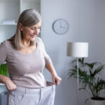 A smiling older woman with gray hair pulls the waistband of her loose-fitting pants, showing noticeable weight loss. She stands in a bright, modern living room.