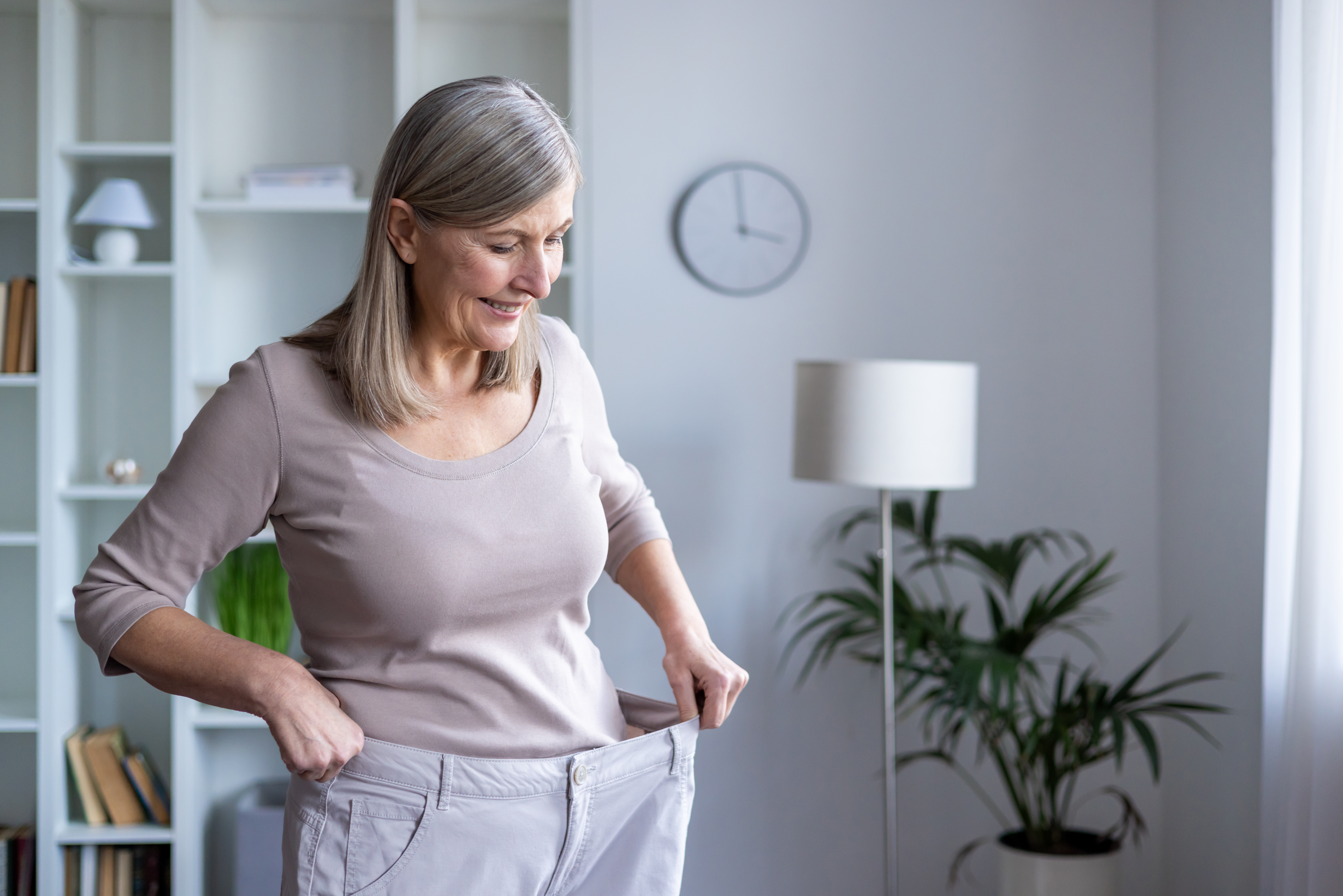 A smiling older woman with gray hair pulls the waistband of her loose-fitting pants, showing noticeable weight loss. She stands in a bright, modern living room.