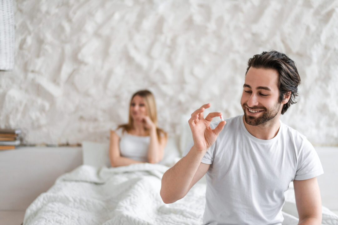 A smiling man in a white t-shirt holds a pill in his hand while sitting on a bed, with a woman in the background looking on.