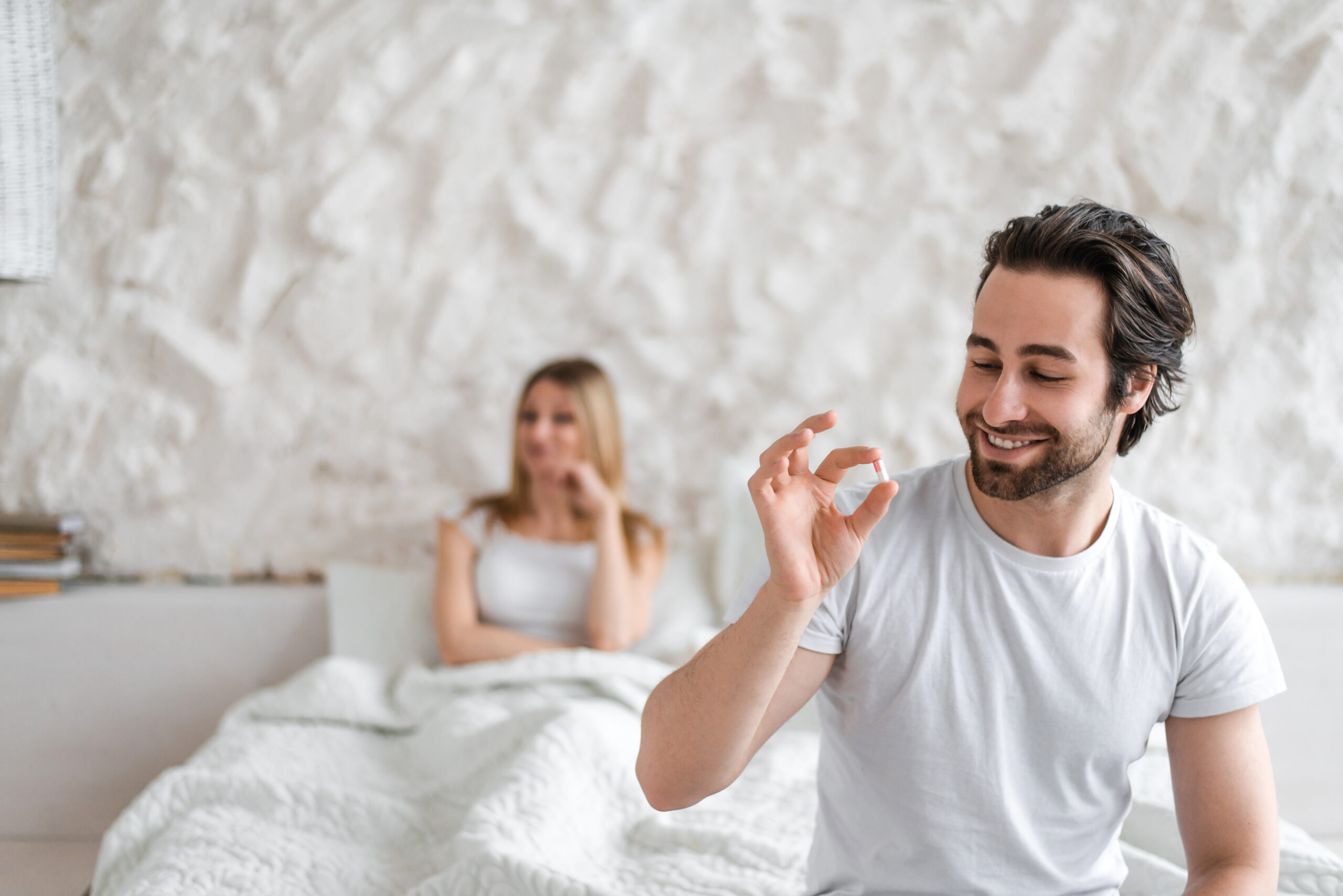 A smiling man in a white t-shirt holds a pill in his hand while sitting on a bed, with a woman in the background looking on.